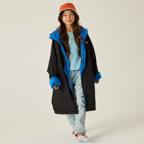 A young girl in a black coat with blue lining and cuffs, light blue shirt, patterned jeans, and orange bucket hat, smiles while walking confidently.