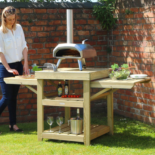 Wooden outdoor pizza oven table with fold-out side shelves and lower storage, shown on a lawn with a pizza oven and food preparation.