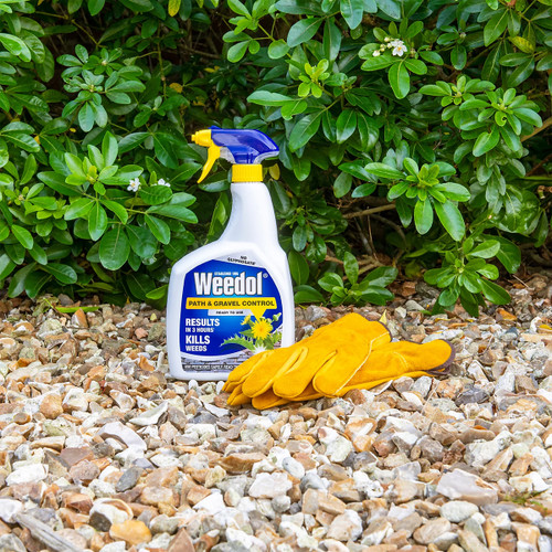A bottle of Weedol weed killer and yellow gloves rest on gravel, with lush green shrubs in the background, suggesting garden maintenance.