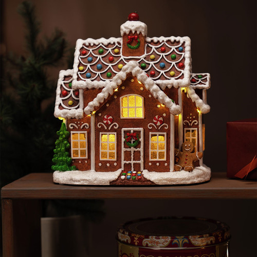 Illuminated gingerbread house Christmas decoration with colourful icing details, snow-covered roof, festive wreaths, gingerbread figure, and warm glowing windows displayed on a wooden side table.