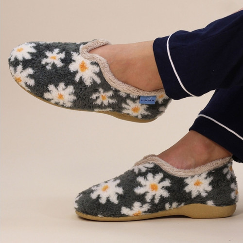 Side view of a pair of women's slippers worn by a person in navy pyjamas, featuring a fluffy charcoal grey upper with white and orange daisy patterns, beige faux fur trim, tan rubber soles, and a blue "LUNAR" brand tag near the collar.