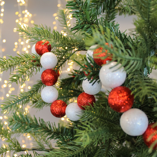 Close-up of a Christmas tree branch with a garland of alternating red and white glittery balls. Soft, warm fairy lights glow in the background. Festive and joyful.