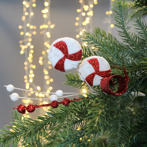 Festive Christmas decoration with glittery red and white peppermint candies and red baubles on a green pine branch against warm fairy lights.
