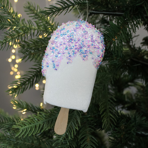 Festive popsicle ornament with pastel sequins and a wooden stick hangs on a Christmas tree branch. Warm lights softly glow in the background.