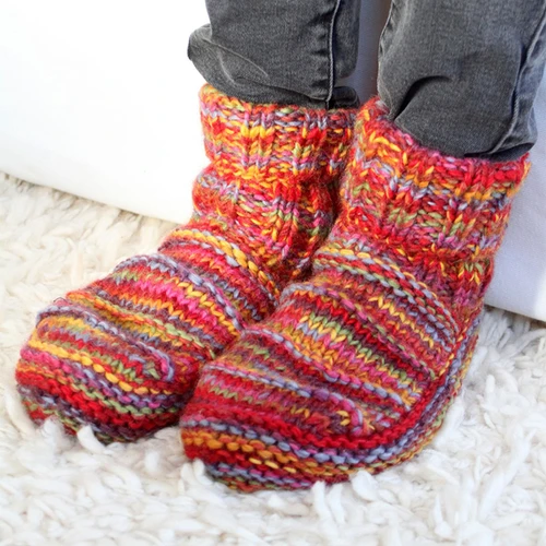 Cosy, multicoloured hand-knitted slippers are worn over dark jeans. The slippers feature vibrant red, orange, and blue hues, set on a plush white carpet.