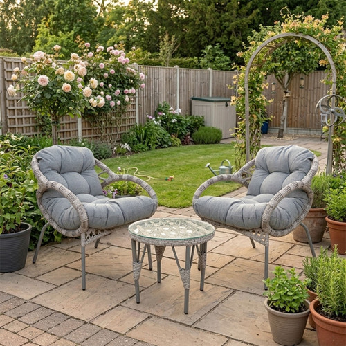 Two grey cushioned wicker chairs with a glass-top table sit on a patio surrounded by potted plants and lush greenery, evoking a serene garden retreat.