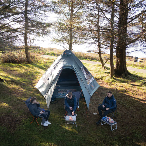 Three people sit on chairs around a small campfire beside a blue tent in a sunlit forest clearing, suggesting a relaxed and peaceful camping scene.