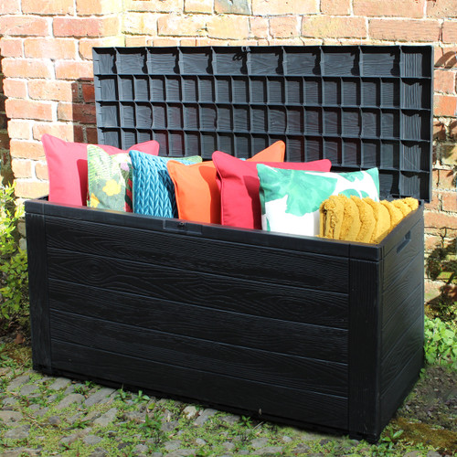 Black wood-effect garden storage box with the lid open, filled with colourful outdoor cushions against a brick wall.