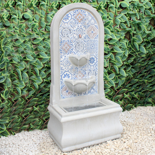 A decorative stone fountain with intricate blue tile patterns stands against lush green foliage, creating a serene and elegant garden atmosphere.