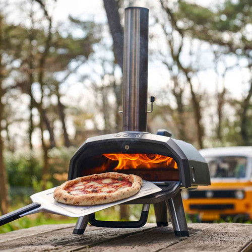 Ooni portable outdoor pizza oven with wood-fired flame cooking a fresh pizza on a metal peel outdoors.