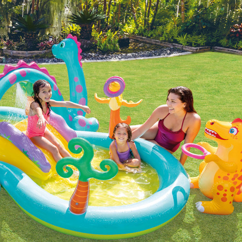 Children playing in a dinosaur-themed inflatable water play centre with slide, sprayer and paddling pool in a garden.