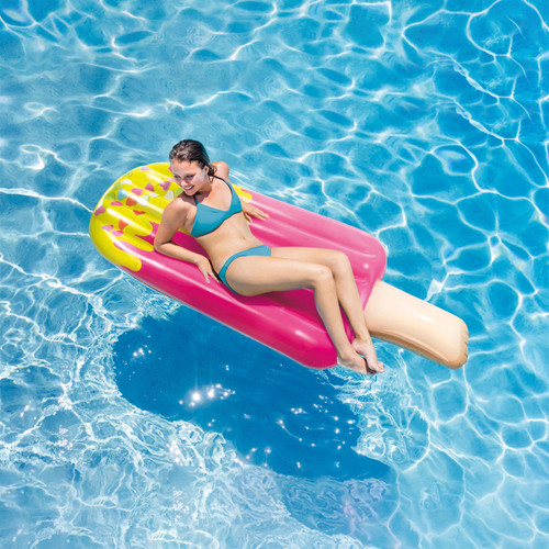 Person relaxing on a pink inflatable popsicle pool float, floating on clear blue water in a swimming pool.
