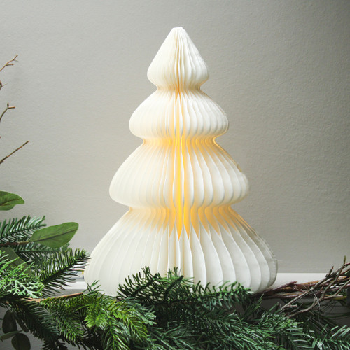 Pre-lit white paper Christmas tree decoration illuminated with a warm white glow, displayed among festive greenery.
