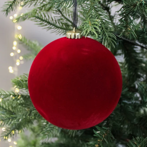 A large red velvet Christmas ornament hangs on a green tree branch with twinkling lights in the background, creating a festive, cozy atmosphere.