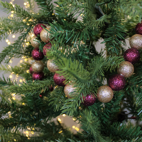 A close-up of a Christmas tree with lush green branches adorned with a string of glittery red and gold baubles. Soft white lights create a warm, festive glow.