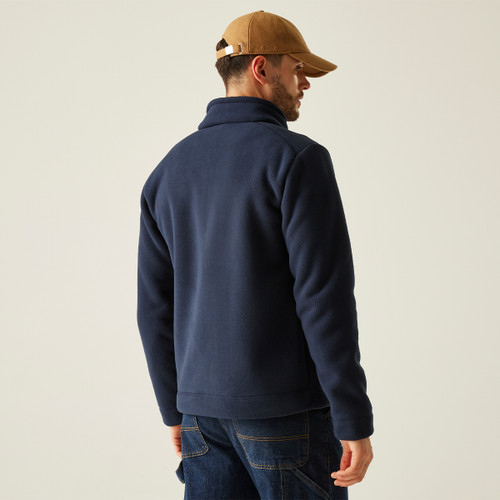 A man wearing a blue fleece jacket and a brown cap stands facing away, set against a plain background. The mood is casual and relaxed.