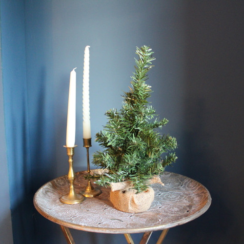 Small artificial tabletop Christmas tree displayed on a decorative table, featuring realistic green pine branches and a rustic burlap-wrapped base.