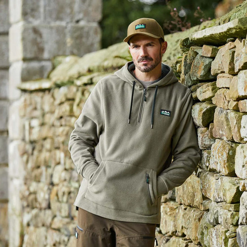 Man wearing a smokey olive Ridgeline hoodie leaning against a dry stone wall outdoors.
