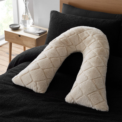 Beige diamond-textured body cushion in a v-shape on a black bedspread with a wooden bedside table and vase.