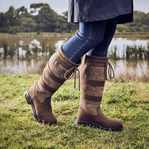 Person in jeans and brown boots walking on grass by a lake.
