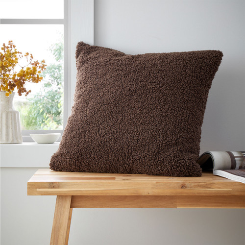 A chocolate brown shaggy cushion on a wooden bench by a window with a vase of flowers.