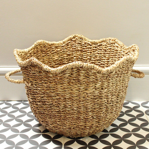 A large scalloped edged seagrass basket with handles on a grey background on a grey background and tiled floor