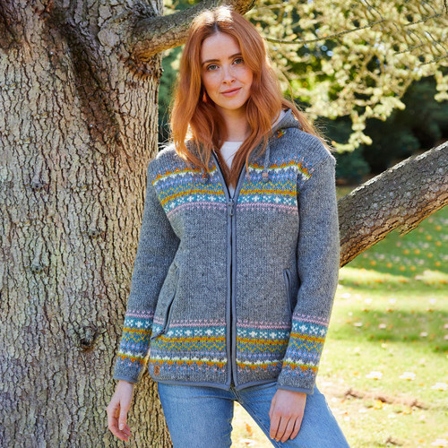 A woman standing under a tree wearing a grey zip up knit hoodie with a multicolour fair isle pattern across the chest and bottom of the jumper.