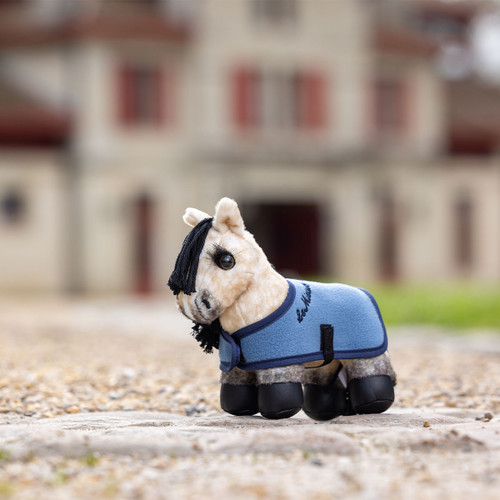 A plush toy horse wearing a blue coat stands on a pebbled path, with a blurred manor house in the background, creating a quaint and whimsical scene.