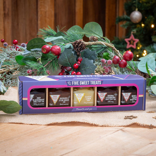 A boxed set of five jam jars sits on a wooden table with festive greenery and red berries in the background. The packaging is colourful and inviting.