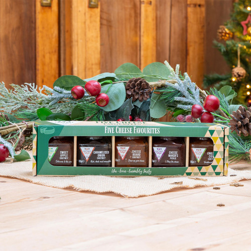 Set of gourmet chutneys labelled "Five Cheese Favourites" on a wooden table with festive greenery and red berries. Christmas tree blurred in the background. Warm, festive ambiance.