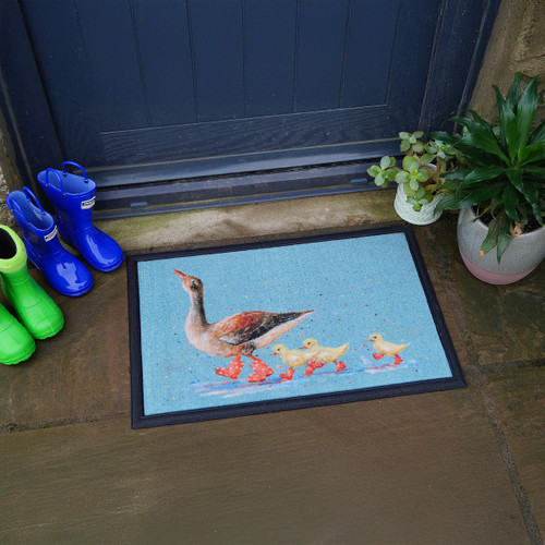 Doormat with a goose and goslings in red boots. Nearby, bright green and blue boots are against a blue door, alongside a potted plant on stone tiles.