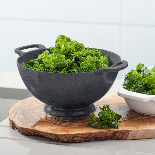 Fresh kale in a dark grey colander with water droplets on a wooden cutting board.