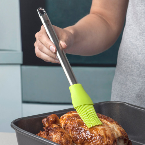A hand holding a green silicone brush, glazing chicken in a roasting pan.