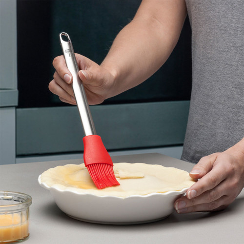 Person brushes egg wash on dough in a white pie dish with a red silicone brush.