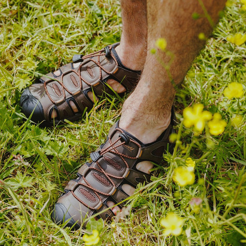 A person wearing dark brown sandals stands on lush, green grass surrounded by yellow wildflowers, conveying a relaxed, nature-inspired vibe.