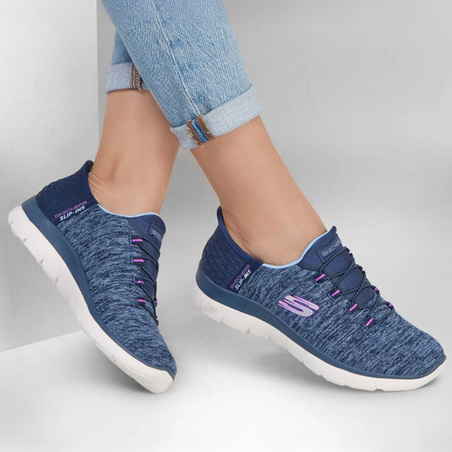 Person wearing light blue jeans and blue slip-on sneakers with pink accents. The shoes have a casual, comfortable look against a grey background.