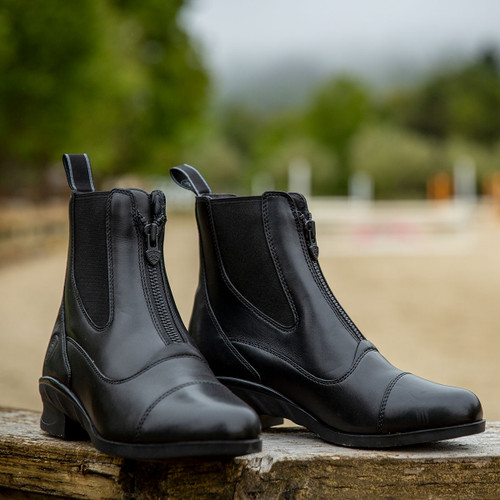 Black leather equestrian boots with front zippers rest on a wooden surface. Blurred green and sandy background suggests an outdoor setting.