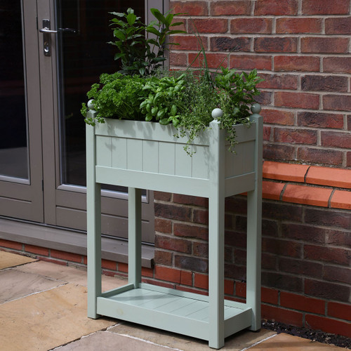 A sage green wooden trough with herbs growing in the deep tray at the top and a shallow tray at the bottom, positioned against an outdoor wall.