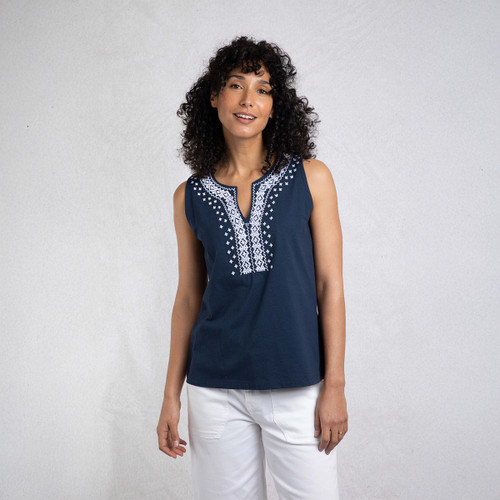 A woman with curly hair poses in a navy sleeveless top featuring white embroidered patterns, paired with white pants against a grey backdrop.