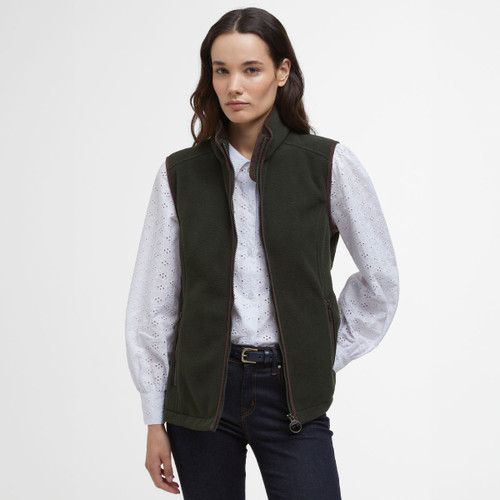 A woman with long hair wears a dark green zip-up vest over a white, long-sleeved blouse with subtle patterns, paired with dark jeans. Neutral expression.