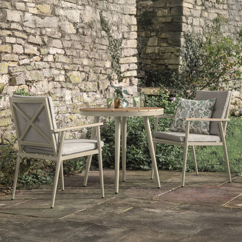 A cosy outdoor seating area featuring two light-coloured chairs and a round wooden table, surrounded by greenery.