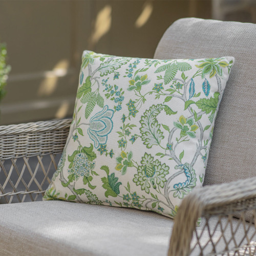 A decorative cushion with vibrant green and blue floral patterns rests on a light-coloured wicker chair, illuminated by soft sunlight.