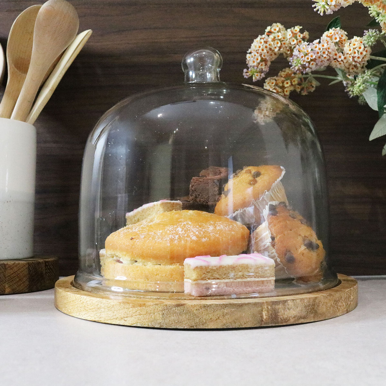 A glass cake dome with a wooden base filled with cakes.