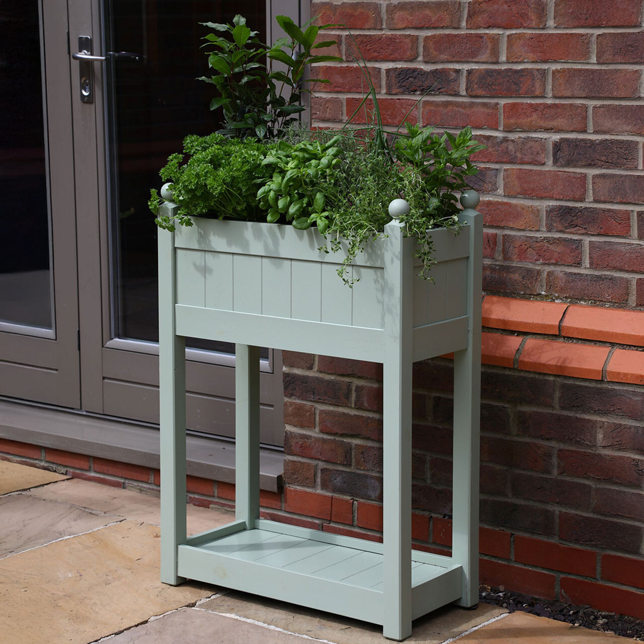 A view of a grey tall planter filled with herbs next to a brick house