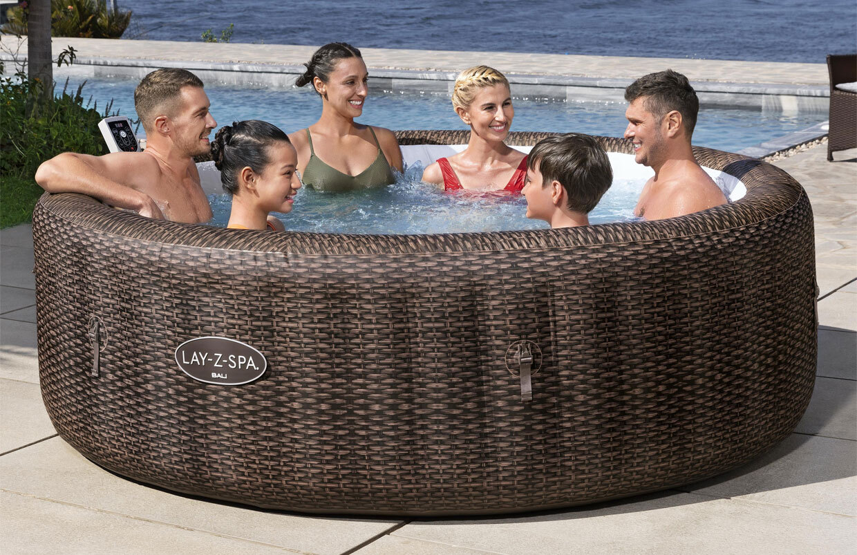 Group of adults and children relaxing together in a Lay-Z-Spa Bali inflatable hot tub with wicker-effect sides, set outdoors beside a swimming pool on a sunny day.