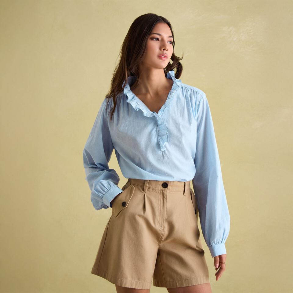 A view of a woman wearing a blue long sleeve V-neck frilly blouse with nude tailored shorts on a beige background 