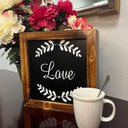 Xtreme Life 4K Wood Frame Hidden Security Camera, decorative wooden frame with the word 'Love' and leaf design on a black background, surrounded by a bouquet of pink and white flowers.
