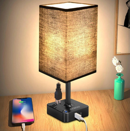 Csc electronics UHD 4K WiFi Lamp USB Charging Station Bedside Security Camera with Live View WiFi + DVR, illuminated lamp on a wooden desk with a smartphone connected via USB, and a small decorative vase with white flowers.