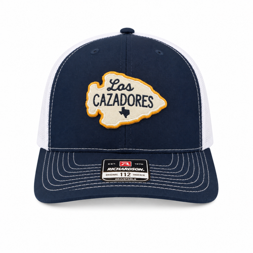 Front view of the Los Cazadores navy and white trucker cap with arrowhead patch, contrast stitching, and brim sticker on a clean white background.