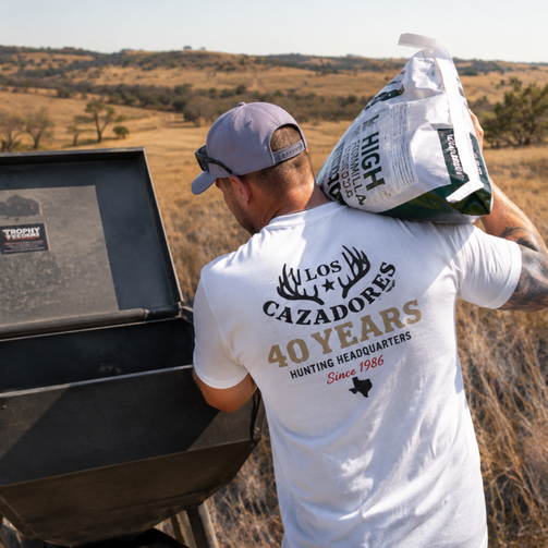 back side of a male model wearing the los cazadores 40 years short sleeve tee while filling a protein feeder in a pasture.